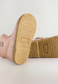 Różowe buty pokazane od dołu, z widoczną fakturowaną podeszwą i złotymi akcentami, na jasnym, neutralnym tle.