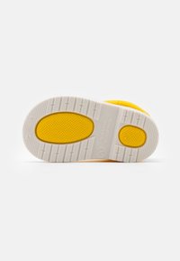 Semelle de sneaker jaune avec un dessous en caoutchouc blanc texturé, comportant des sections de grip à motifs et un accent circulaire près du talon.