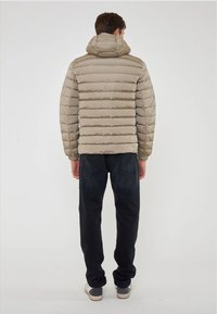 Giacca puffer beige con cuciture verticali e cappuccio, abbinata a jeans scuri e sneaker grigie, vista da dietro.