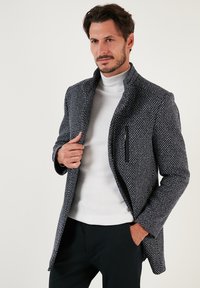 Cappotto strutturato blu navy e bianco con colletto alto, dotato di una tasca laterale con zip. Indossato sopra un dolcevita bianco e pantaloni neri.