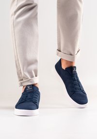 Marineblaue Strick-Sneaker mit Schnürdesign, weißen Gummisohlen und markiertem Zunge. Kombiniert mit gekrempelten hellbeigen Hosen, die den Knöchel zeigen.