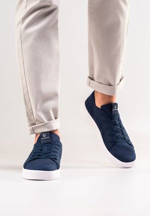 Navy knit sneakers with lace-up design, white rubber soles, and branded tongue. Worn with rolled light beige pants showcasing the ankle.