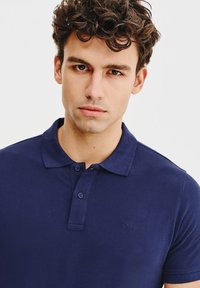Granatowa koszulka polo wykonana z bawełny, z klasycznym kołnierzykiem, dwoma guzikami i subtelnym logo marki na piersi. Gładka faktura.