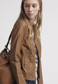 Bruine leren jas met een nauwsluitend ontwerp, voorrits, zijzakken en een gestructureerde afwerking, gedragen met een patroon top en een bruine tas.