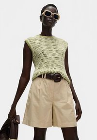 Jasnozielony dzianinowy top bez rękawów, beżowe plisowane szorty, brązowy pasek, trzyma brązową kurtkę, nosi duże beżowe okulary przeciwsłoneczne.