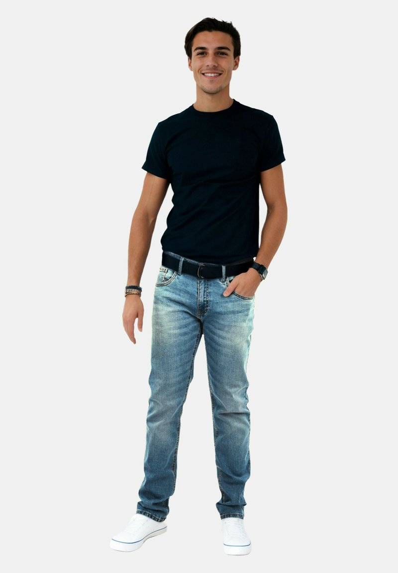 Young man standing, smiling, wearing a black t-shirt, blue jeans, white sneakers, and wrist accessories against a plain white background.