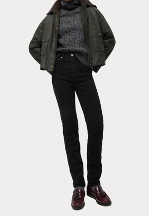 Olivgrüne Bomberjacke über einem grauen Grobstrickpullover, kombiniert mit hochgeschnittenen schwarzen Jeans und burgunderfarbenen Slippern mit Fransen.