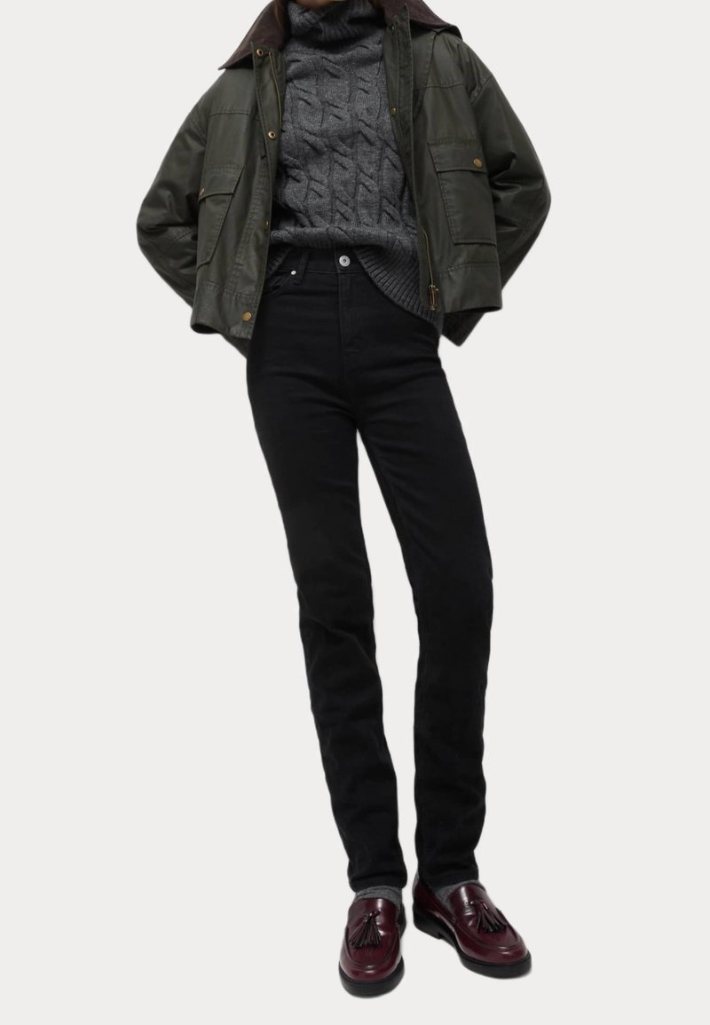 Olivově zelená bomber bunda přes šedý pletený svetr, doplněná o vysoké černé džíny a burgundské mokasíny s třásněmi.