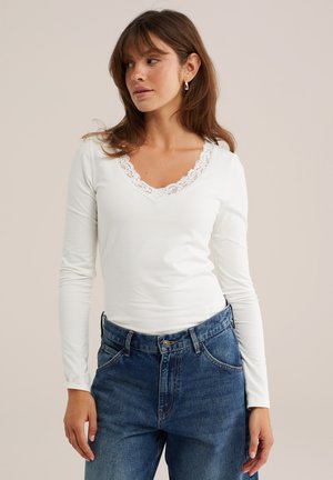 Femme aux cheveux bruns portant un haut blanc à manches longues avec bordure en dentelle et un jean bleu, regardant légèrement vers sa gauche sur un fond uni.