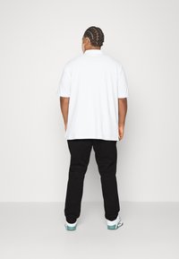 Hvit kortermet polo-shirt bæres med svarte bukser, som viser glatt stoff og en avslappet passform. Synlig merking og kontrasterende detaljer på skoene.
