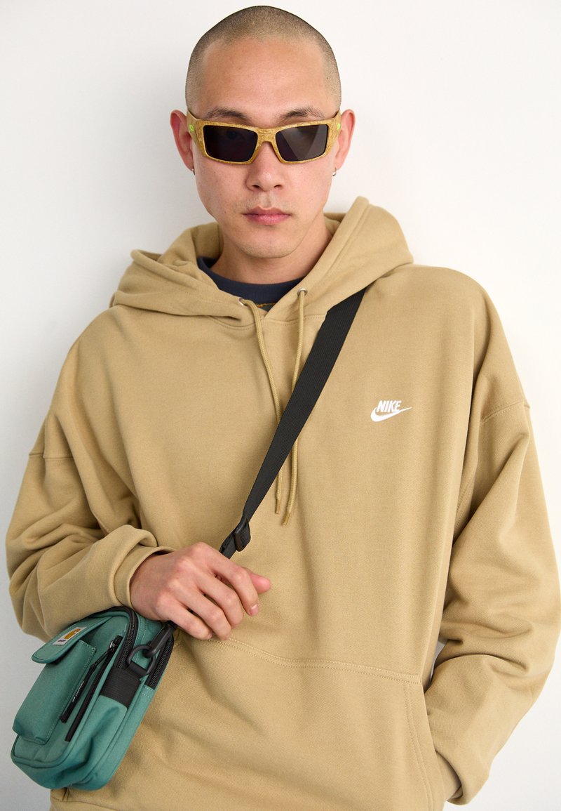 Haut à capuche beige de Nike avec cordon de serrage. Sac bandoulière noir à la main, couleur teal avec fermeture éclair. Lunettes de soleil sombres avec un design de cadre unique.