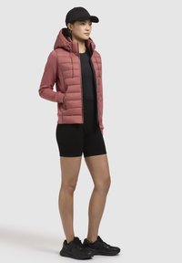 Lichtroze gewatteerde jas met een capuchon, zwarte aansluitende top en zwarte shorts. Gekozen met zwarte sportschoenen en een zwarte pet.