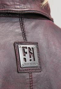 Vinröd läderjacka med texturerad yta, metallpatch med utskurna bokstäver "FN" och synliga sömdetaljer längs sömmen.