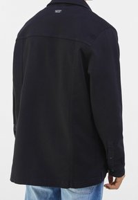 Zwarte katoenen jas met een relaxte pasvorm, voorzien van een knoopmanchet, subtiele stiksels en een logotag op de achterkant van de kraag.