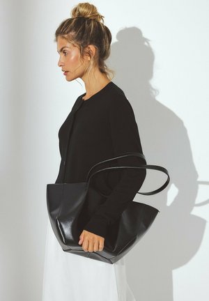 Sac fourre-tout en cuir noir avec une texture lisse, une large ouverture et une forme angulaire distincte. Dispose de deux poignées allongées pour le transport.
