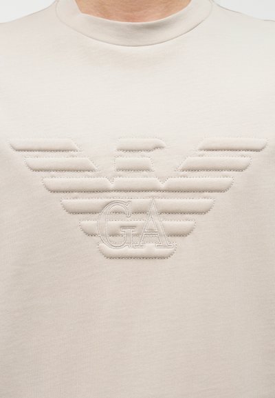 Κοντινό πλάνο ενός ανοιχτό μπεζ πουκαμίσου με ραμμένο έμβλημα με φτερά και τα γράμματα "GA" στο κέντρο του στήθους.