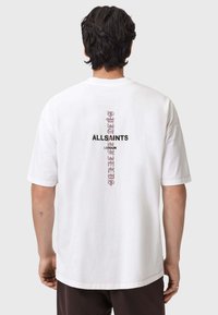 Valkoinen puuvillateepaita, jossa on musta ja violetti pystysuora graafinen kuvio selässä, jossa lukee "ALLSAINTS" ja "LONDON". Lyhyet hihat.