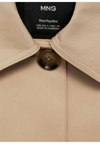 Col de manteau beige déperlant avec un bouton marron foncé et une étiquette intérieure indiquant la marque, la taille et les détails d'origine.