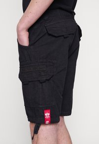 Personne portant un short cargo noir avec plusieurs poches et un patch logo Alpha Industries rouge près de l'ourlet sur fond blanc.