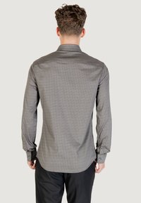 Uomo con capelli corti e ricci, che indossa una camicia a maniche lunghe grigia e a fantasia con pantaloni neri, in piedi di spalle contro uno sfondo neutro.