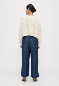 Woman with dark hair in a bun wearing large hoop earrings, a cream cable-knit cropped sweater, wide-leg dark blue jeans, and taupe heeled boots.