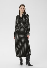 Fekete pöttyös ruha hosszú ujjakkal és gombos gallérral. Hosszú, áramló kialakítás jellemzi, és térdig érő fekete csizmával kombinálható.