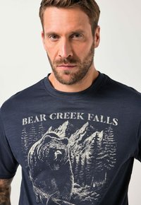 Námořnická bavlněná tričko s opotřebovaným grafickým motivem medvěda a horské scenérie v bílé barvě, kulatým výstřihem a krátkými rukávy.