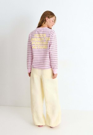 Mujer con camisa de rayas rosas de manga larga con el texto "Enfoca la energía en acciones positivas" en la espalda, combinada con pantalones anchos de color crema.