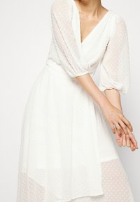 Vit klänning med ett texturerat prickigt mönster, V-ringning och puffärmar. Kjolen är lagrad, vilket skapar en flödesform.