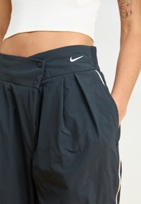 Črne najlon hlače z nabranim dizajnom, dvojno gumbno zapiranje, stranskimi žepi in belim Nike logotipom blizu pasu.