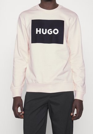 Homme portant un sweat-shirt rose clair avec un logo rectangulaire noir et le texte blanc « HUGO », associé à un pantalon noir, debout sur fond blanc.