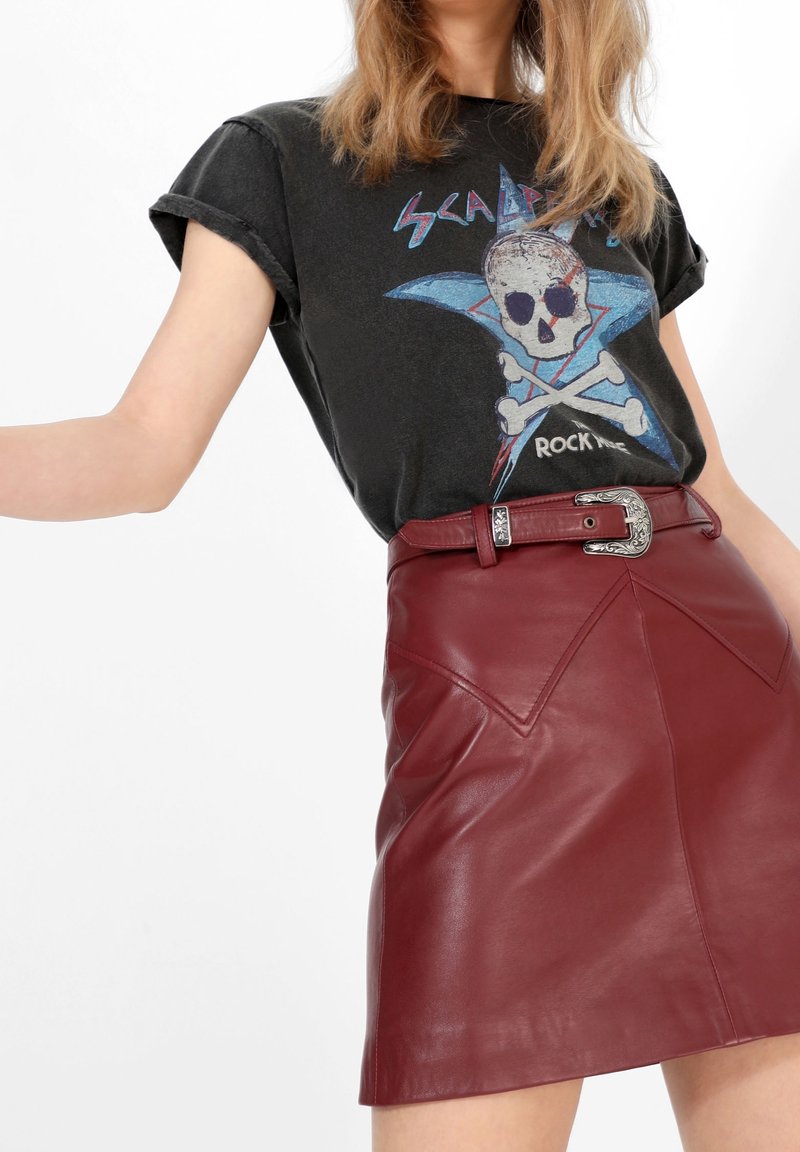 Femme portant un t-shirt noir à motif crâne et une jupe en cuir rouge taille haute avec une ceinture décorative à boucle argentée.