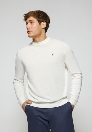 Suéter blanco de cuello redondo hecho de un tejido suave, con mangas largas y un pequeño logotipo azul marino en el pecho. Combinado con pantalones azul oscuro.