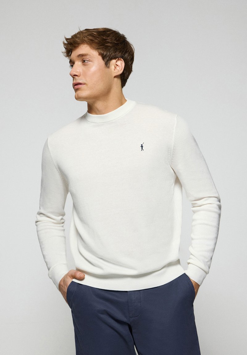 Maglione bianco a collo rotondo realizzato in un tessuto morbido, con maniche lunghe e un piccolo logo blu navy sul petto. Indossato con pantaloni blu scuro.