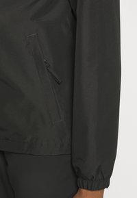 Svart lättviktsjacka med en slät yta, elastiska ärmslut och en ficka med dragkedja på sidan. Enkelt design utan mönster eller detaljer.