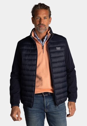 Man draagt een marineblauw doorgestikt jack over een perzikkleurige trui met kwart-ritssluiting en blauwe spijkerbroek, staand tegen een effen lichte achtergrond.