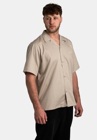 Chemise beige à manches courtes, avec un col décontracté, fabriquée dans un tissu lisse, présentant un logo discret sur le côté gauche de la poitrine.