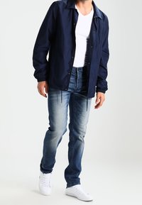 Chaqueta ligera navy con cuello, cierre de botones, puños elásticos; combinada con jeans azul desgastado y zapatillas blancas.