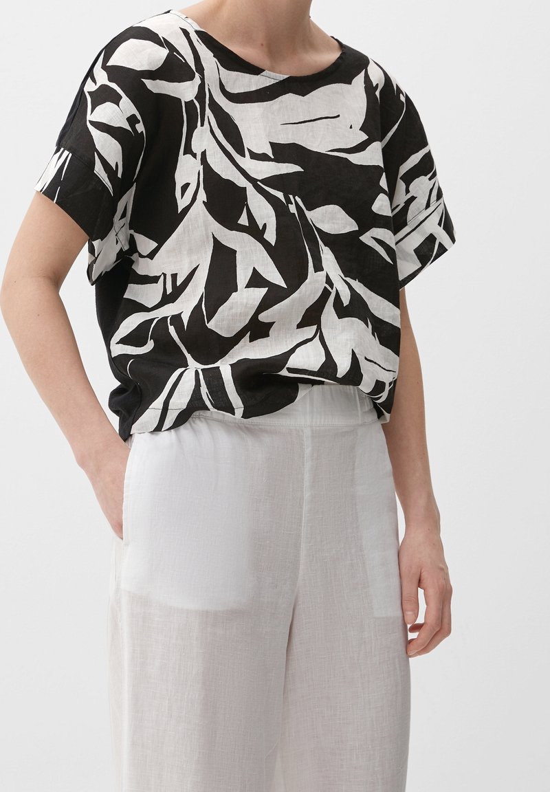 Personne portant un haut ample à manches courtes avec un imprimé abstrait noir et blanc, rentré dans un pantalon en lin blanc taille haute avec poches latérales.