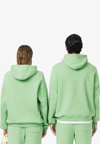 Felpe verdi realizzate in tessuto morbido, con una vestibilità comoda, polsini a coste e tasche a marsupio. Entrambi i modelli indossano pantaloni coordinati.