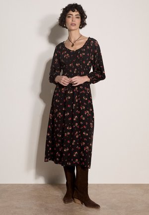 Frau mit kurzem lockigem Haar, die ein schwarzes florales Midikleid und braune Stiefel trägt, steht vor einem schlichten hellen Hintergrund.