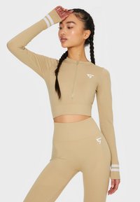 Beige Sport-Crop-Top mit langen Ärmeln, viertelzip vorne und weißen Streifenakzenten; kombiniert mit hoch taillierten passenden Leggings.