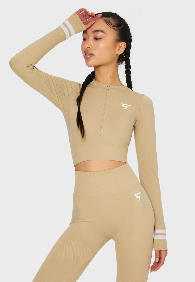 Beige Sport-Crop-Top mit langen Ärmeln, viertelzip vorne und weißen Streifenakzenten; kombiniert mit hoch taillierten passenden Leggings.