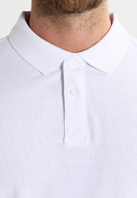 White polo shirt made from textured fabric. Features a collar, three-button placket, and a triangular stitching detail on the hem.
