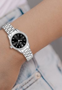 Sudraba pulkstenis ar teksturētu melnu ciparnīcu, sudraba stundu rādītājiem un iegrimušu bezel. Aprīkots ar nerūsējošā tērauda aproci uz rokas.