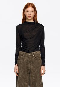 Czarna bluzka z długim rękawem z przezroczystej tkaniny z asymetrycznym drapowaniem. Połączona z oliwkowozielonymi szerokimi spodniami o fakturowanym wzorze.