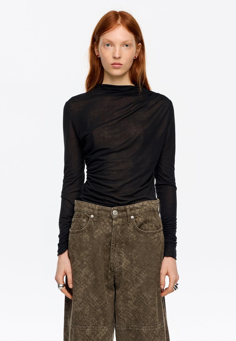 Czarna bluzka z długim rękawem z przezroczystej tkaniny z asymetrycznym drapowaniem. Połączona z oliwkowozielonymi szerokimi spodniami o fakturowanym wzorze.
