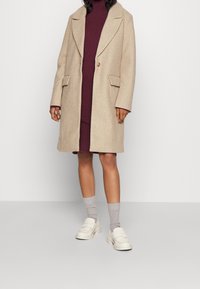 Abrigo de lana beige con un único botón, cuello con muesca y dos bolsillos frontales. Llevado sobre un vestido burdeos de canalé, combinado con calcetas grises y mocasines blancos.