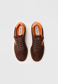 Sapatilhas de camurça castanha com detalhes laranja, design com atacadores, língua texturizada, sola flexível e detalhe do logotipo na lateral.