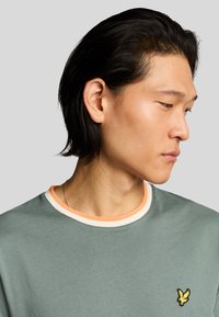Grøn crew-neck t-shirt med en kontrasterende orange og hvid ribbet krave, med et lille gult broderet logo på brystet.